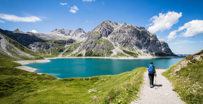 Eine einzelne Person mit Rucksack wandert auf einem schmalen Kiesweg entlang eines türkisblauen Sees, umgeben von majestätischen, grauen Berggipfeln. Ein klarer Himmel mit wenigen Wolken dominiert die Szene.