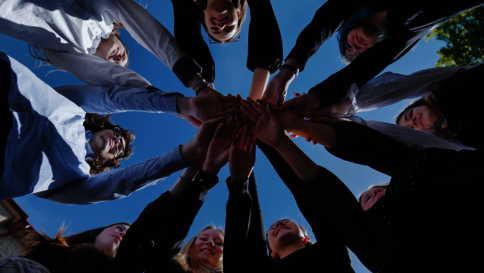 Eine Gruppe von Menschen steht im Kreis und legt ihre Hände gemeinsam in die Mitte übereinander – Symbol für Zusammenarbeit und Gemeinschaft vor blauem Himmel.