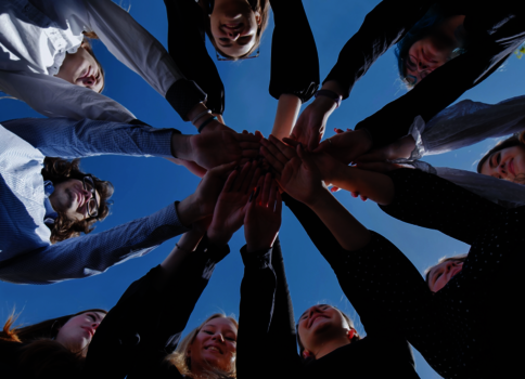 Eine Gruppe von Menschen steht im Kreis und legt ihre Hände gemeinsam in die Mitte übereinander – Symbol für Zusammenarbeit und Gemeinschaft vor blauem Himmel.
