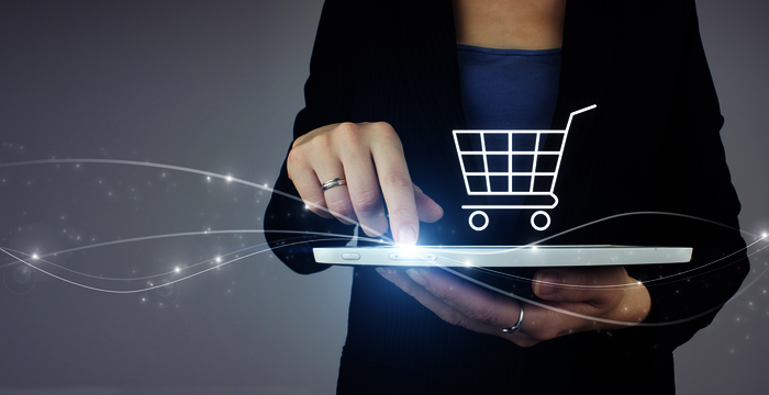 Eine Person in dunkler Kleidung hält ein Tablet; eine Hand tippt darauf, während ein leuchtender Einkaufswagen über dem Tablet erscheint und Online-Shopping symbolisiert.