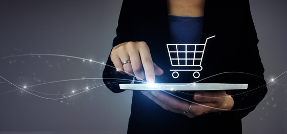 Eine Person in dunkler Kleidung hält ein Tablet; eine Hand tippt darauf, während ein leuchtender Einkaufswagen über dem Tablet erscheint und Online-Shopping symbolisiert.