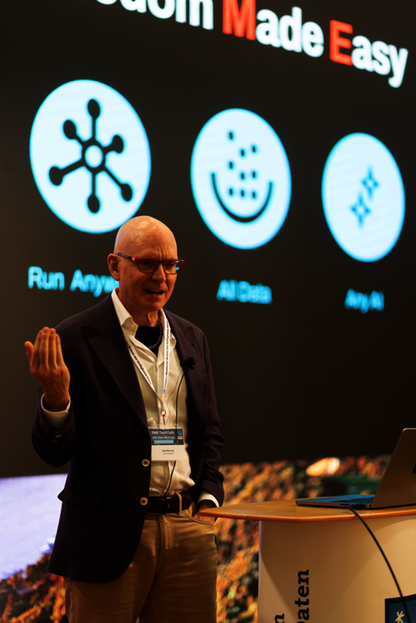 Ein Mann in Blazer mit Namensschild spricht auf einer Bühne, daneben ein Laptop auf dem Pult. Im Hintergrund eine große Projektion mit mehreren blauen Symbolen in Kreisen und einem Satz oben, vermutlich ein Tech-Themen-Panel.