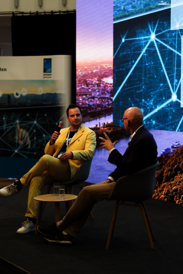 Zwei Männer sitzen auf einer Bühne in Sessel im Gespräch. Der Mann links trägt einen gelben Anzug und hält ein Mikrofon, der Mann rechts in dunkler Jacke gestikuliert. Zwischen ihnen steht ein runder Tisch mit Wasser. Hinter ihnen leuchten digitale Panels mit Stadt- und Netzwerksymbolen.
