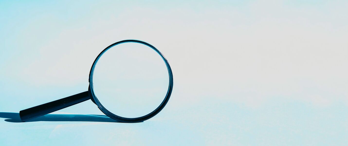 A black-handled magnifying glass rests on a pale blue gradient background, with its circular lens facing upward and casting a long shadow.