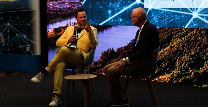 Zwei Männer sitzen auf einer Bühne im Gespräch während eines TechTalk-Events. Links spricht der in Gelb gekleidete Moderator mit Mikrofon, rechts hört ein zweiter Mann im Anzug zu. Im Hintergrund zeigt eine große Bildschirmprojektion Event-Details und ein Landschaftsbild.