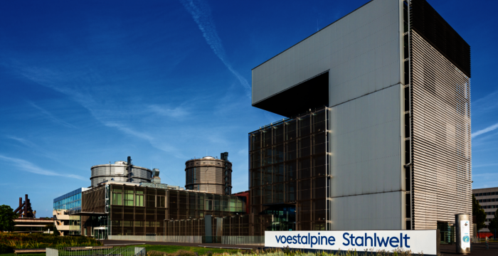 Moderne Industrieanlage mit großem, rechteckigem Hauptgebäude und dunkler Fassadenseite; vorne befindet sich ein Schild mit 'voestalpine Stahlwelt'. Klarer blauer Himmel, Grünflächen und weitere Gebäude im Hintergrund.
