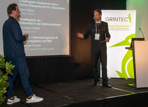 Zwei Männer stehen auf einer Konferenzbühne: Der rechts stehende Sprecher gestikuliert, der links zuhörende Mann schaut zu. Im Hintergrund läuft eine Folienpräsentation, daneben ein Banner mit Logo und ein Rednerpult.