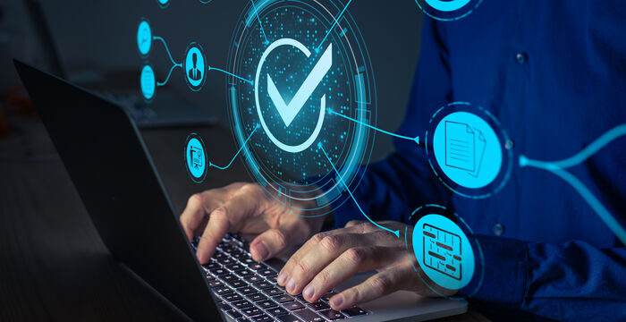Eine Person tippt auf einem Laptop; vor dem Bildschirm schwebende blaue Hologramme zeigen ein zentrales Häkchen, das digitale Verifizierung oder sichere Online-Transaktionen symbolisiert.