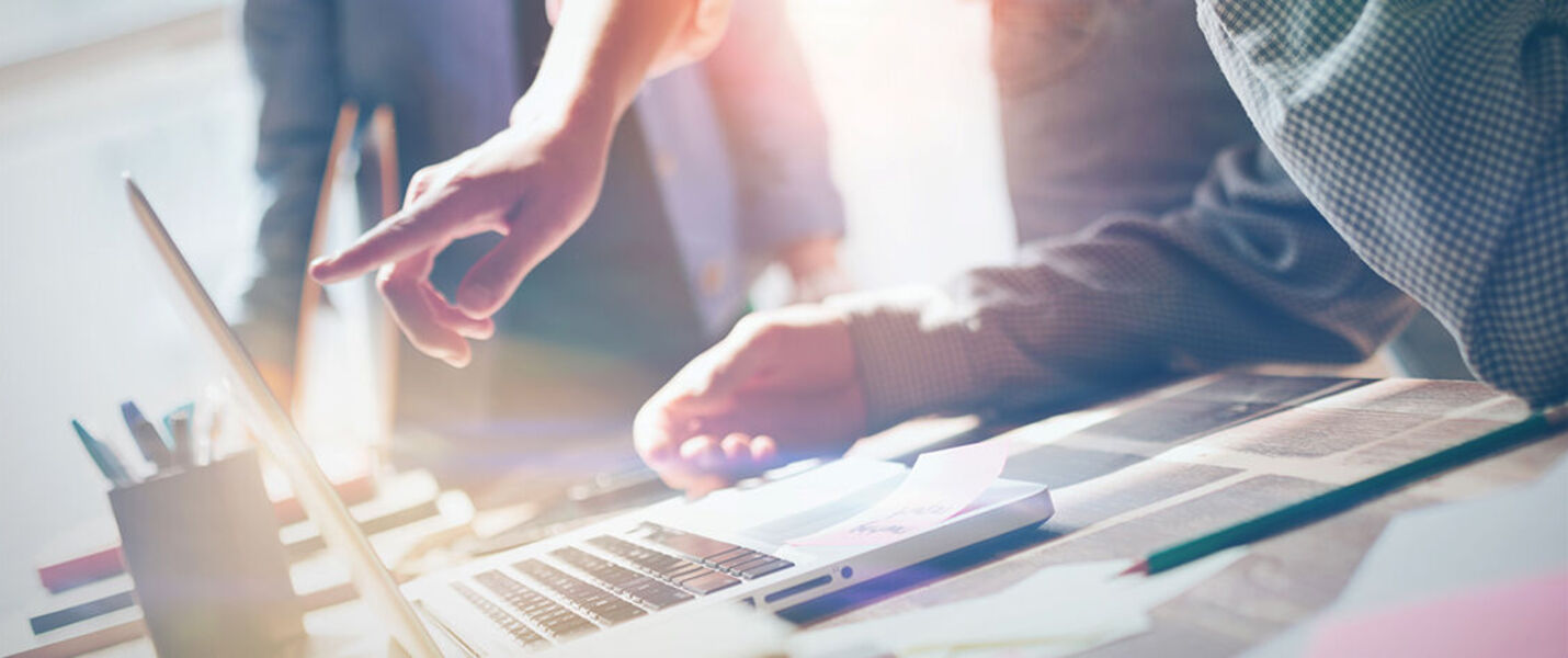 Eine Gruppe von Menschen arbeitet an einem Tisch. Einer zeigt auf das Laptop-Display, während die anderen Notizen und Unterlagen beisteuern. Die Szene vermittelt Zusammenarbeit, Ideenfindung und Fokus in einem hellen Büroraum.