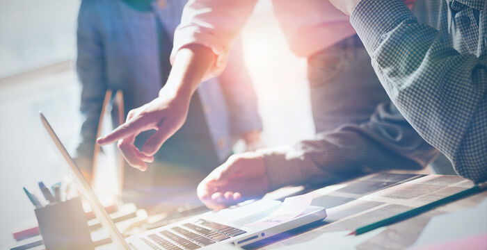 Group of coworkers gathered around a laptop in a bright office, one person pointing at the screen as others watch, with papers and notebooks spread on the desk.
