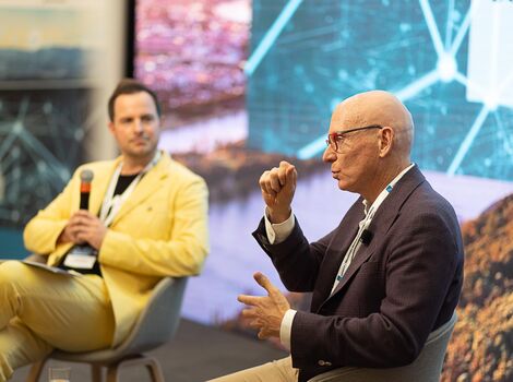 Zwei Männer führen ein Panelgespräch auf einer Bühne. Der linke Mann in gelbem Anzug hält ein Mikrofon; der rechte Mann in dunklem Sakko und Brille spricht mit Handgesten. Im Hintergrund sind digitale Grafiken zu sehen.