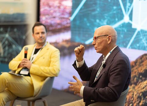 Zwei Männer führen ein Panelgespräch auf einer Bühne. Der linke Mann in gelbem Anzug hält ein Mikrofon; der rechte Mann in dunklem Sakko und Brille spricht mit Handgesten. Im Hintergrund sind digitale Grafiken zu sehen.