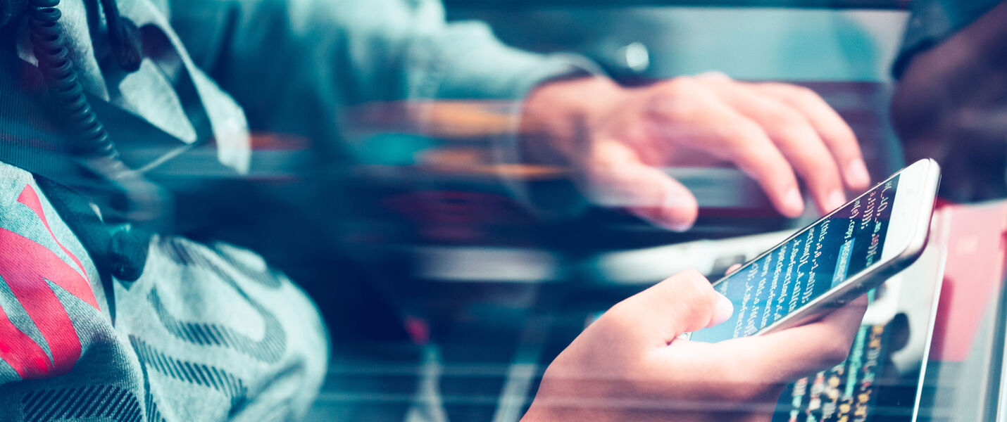 Eine Person hält ein Smartphone in der Hand und tippt darauf; im Hintergrund arbeitet jemand an einer Tastatur.