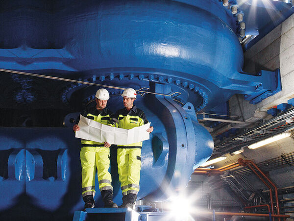 Zwei Arbeiter in leuchtend gelben Schutzanzügen und Helmen stehen auf einer Plattform und betrachten eine große Bauzeichnung. Hinter ihnen dominiert eine riesige blaue Industriemaschine in einer Fabrikhalle.